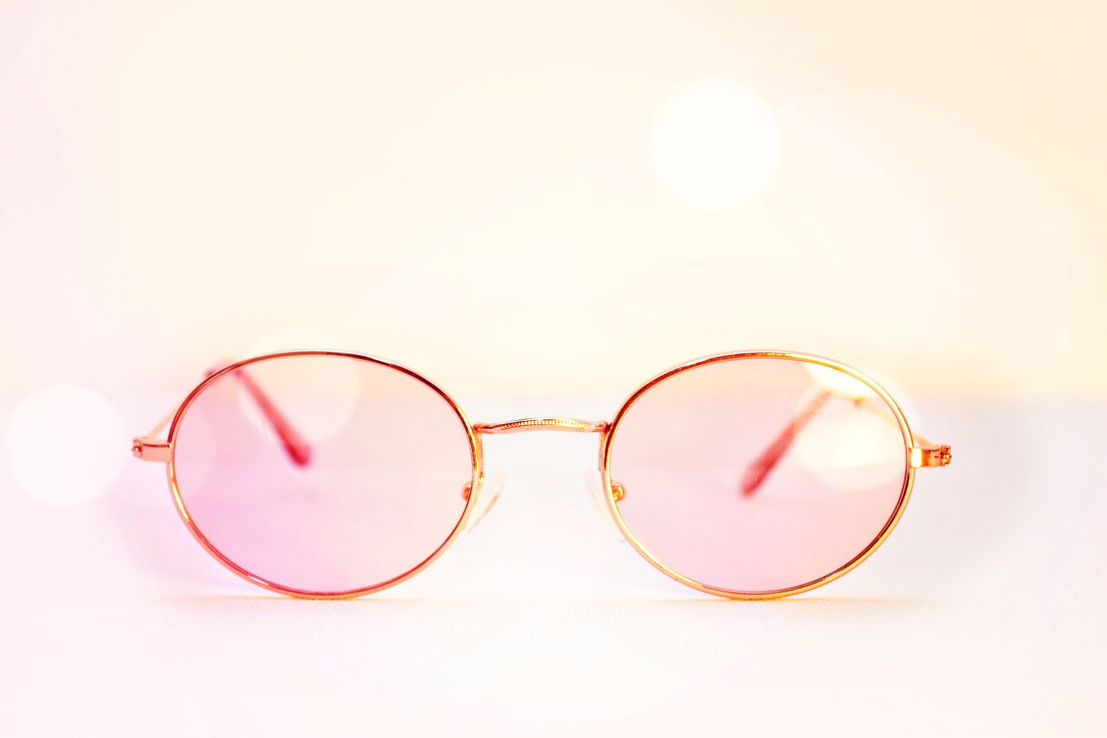 Armação de aro fino em ouro com lentes rosadas claras, sobre fundo claro suave.