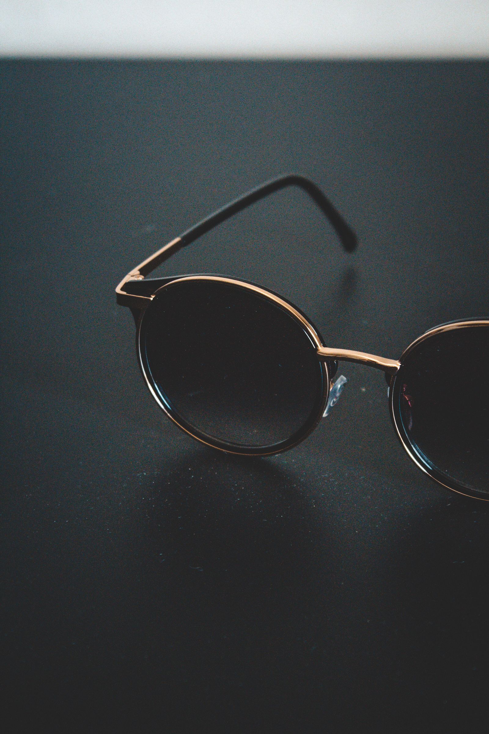 Óculos de sol redondo com aro dourado fino sobre superfície escura, composição minimalista.